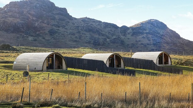 Three rustic, cylindrical cabins with wooden doors, set in a grassy field with a mountainous backdrop. - A good sized, dog friendly pod with silent surroundings. (Archaracle)