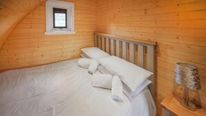 A neatly made bed with white linens and pillows, a wooden headboard, a small window, and a wooden nightstand with a lamp. - A good sized, dog friendly pod with silent surroundings. (Archaracle)