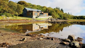 A modern house with a stone foundation, large windows, and a metal roof, situated by a calm lake with a rocky shoreline.