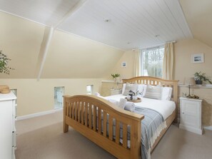 Een slaapkamer met een schuine kap, een houten bed, een nachtkastje met een lamp, een raam met gordijnen, en een klein plankje met planten.