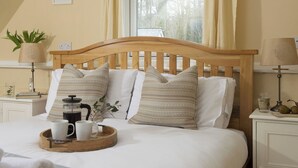A neatly made bed with a wooden headboard, a bedside table with a lamp, a tray with a mug and a coffee pot, and a window with curtains. - 3 bedroom accommodation in Bovey Tracey (Bovey Tracey)