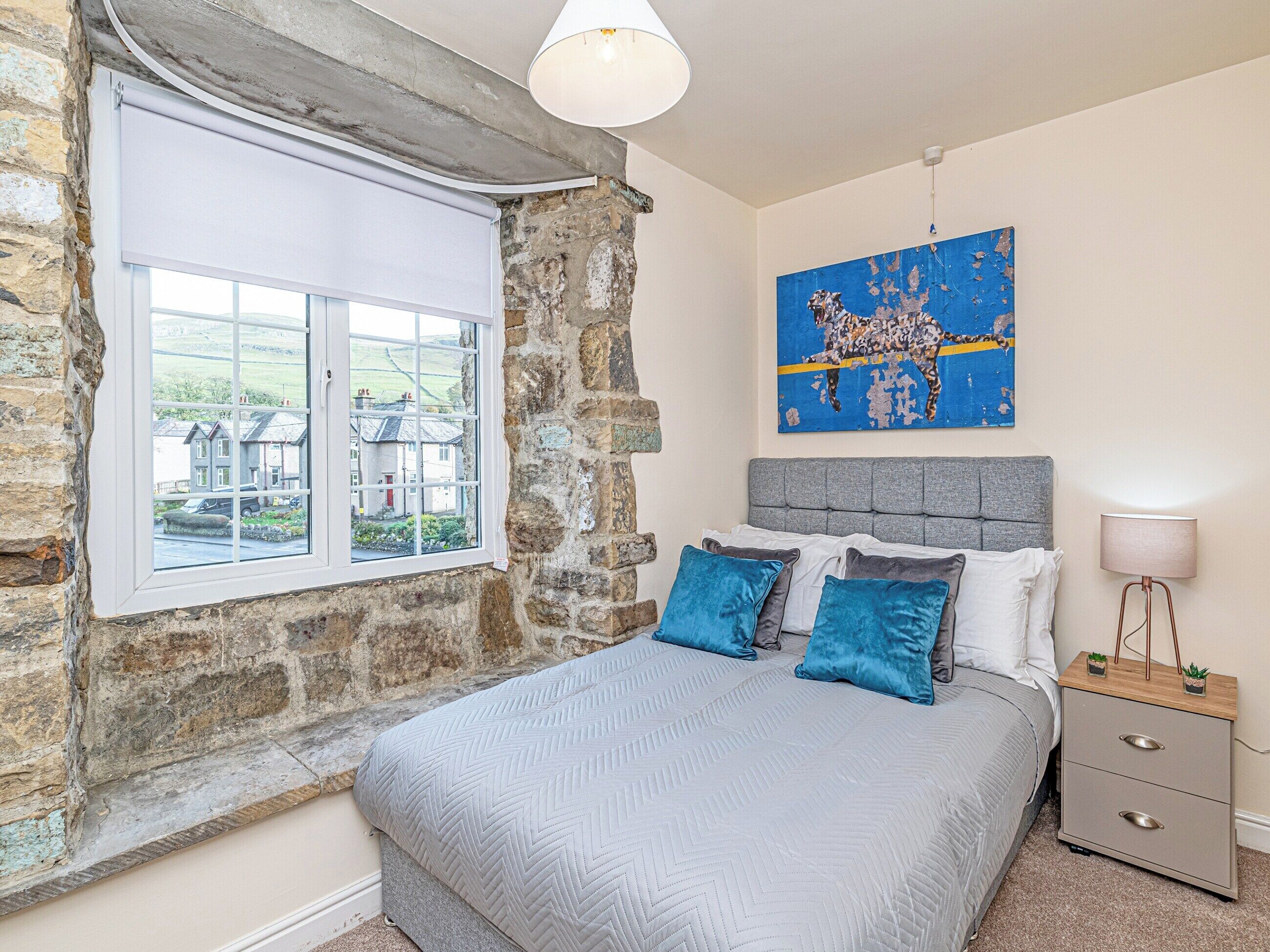 A bedroom with a stone wall, a large window, a bed with blue pillows, a grey headboard, a bedside table with a lamp, and a painting on the wall.