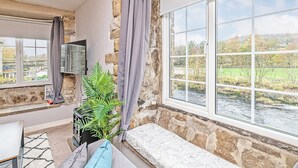 Een gezellige kamer met een stenen muur, een groot raam met uitzicht op een rivier en groen, een bankje en een aan de muur bevestigde televisie.