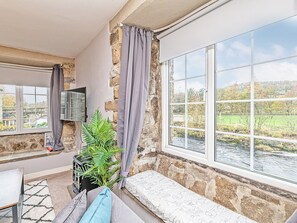 Ein gemütliches Zimmer mit Steinwand, einem großen Fenster mit Blick auf einen Fluss und viel Grün, einer Bank und einem an der Wand montierten Fernseher.