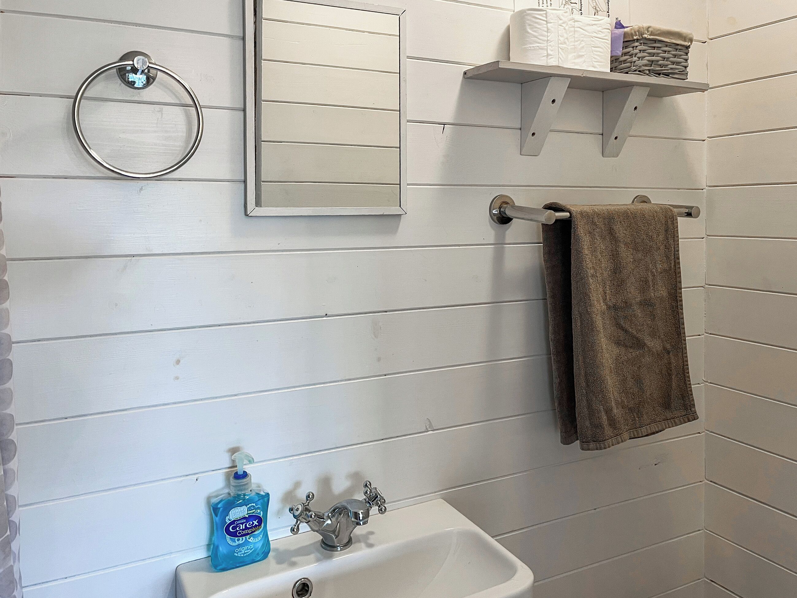 Ein Badezimmer mit einem weißen Waschbecken, einem Spiegel, einem Handtuchhalter, einer Seifenspender und einem Regal mit Pflegeprodukten.