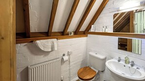 Une petite salle de bain avec un plancher en bois, des murs en briques blanches, un radiateur blanc, des toilettes avec un abattant en bois, un lavabo blanc avec un miroir au-dessus, et une petite poubelle.