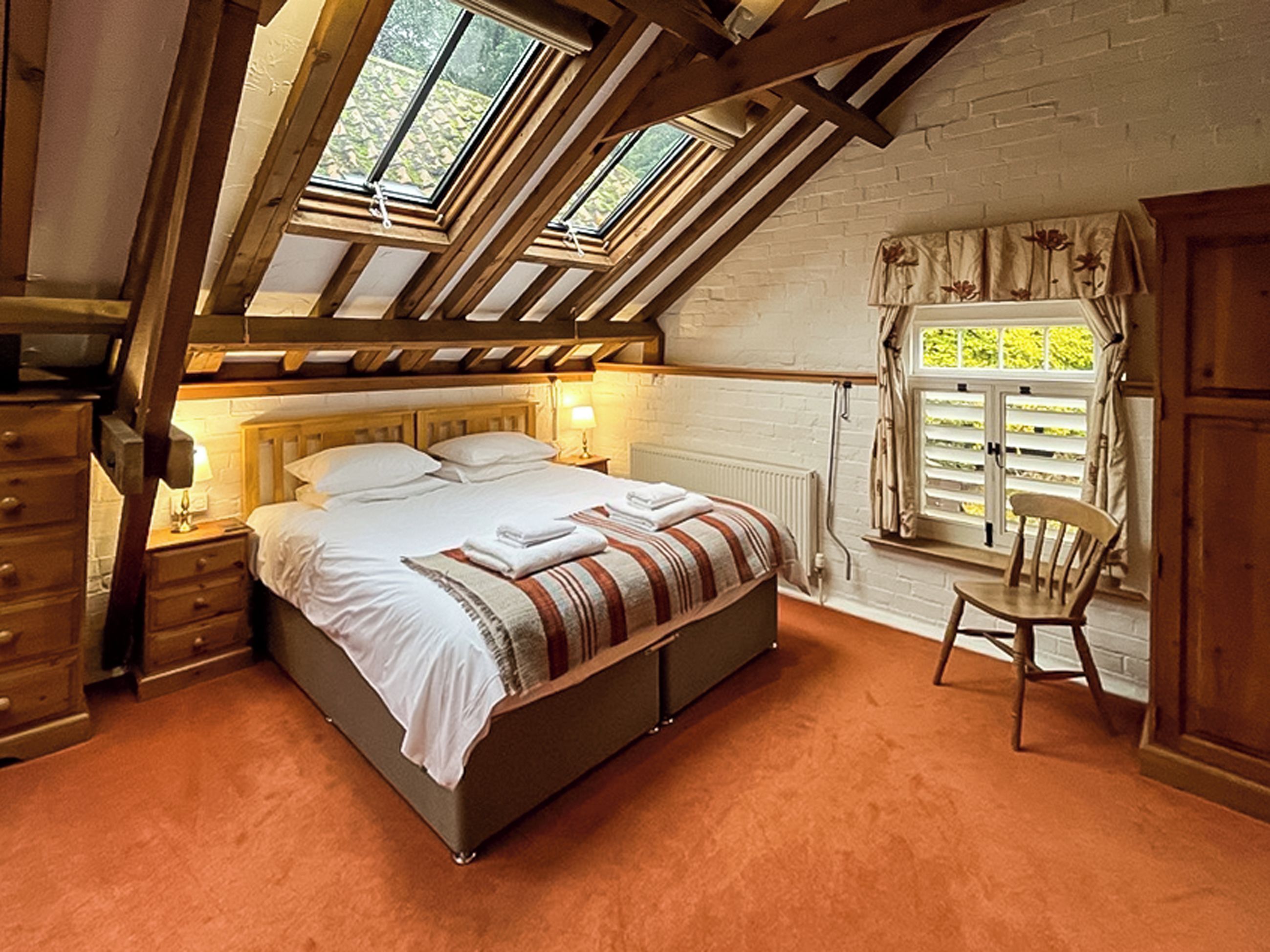 A bedroom with a sloped ceiling, a bed with white linens, a wooden chair, and a window with curtains.