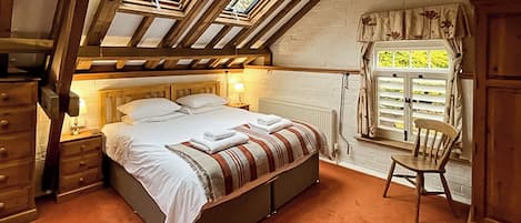 Un dormitorio con techo inclinado, una cama con ropa de cama blanca, una silla de madera y un ventanal con cortinas.