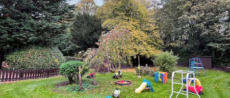 A well-maintained grassy area with a playset, including a slide and a swing set, surrounded by trees and a wooden fence.
