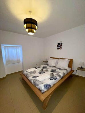 A bedroom with a bed, a bedside table, a hanging light fixture, a window with curtains, and a wall-mounted decoration.