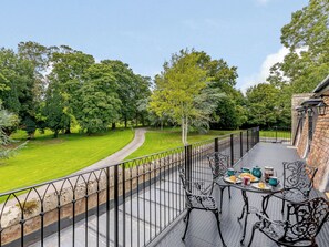 Un balcón con una mesa dispuesta para el desayuno, con vistas a un césped cuidado y árboles.