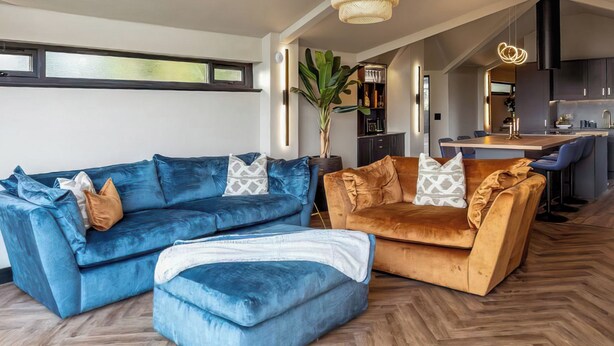 Een moderne woonkamer met een visgraat houten vloer, een blauwe hoekbank, een bruine bank en een eethoek, met een keuken op de achtergrond.