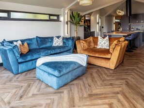 Un salon moderne avec un parquet en chevrons, un canapé d’angle bleu, un canapé marron et un coin repas avec une cuisine en arrière-plan.