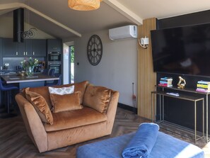 A living room with a sofa, a TV, and a dining area with a kitchen in the background. - Deerview Stables - UK51043 (Bonvilston)