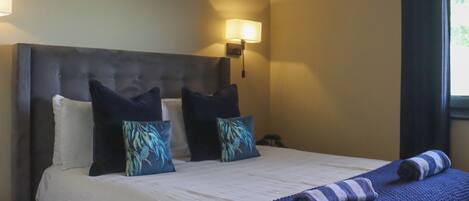 A hotel room with a large bed, two pillows, a headboard, a wall-mounted light, and a window with curtains.