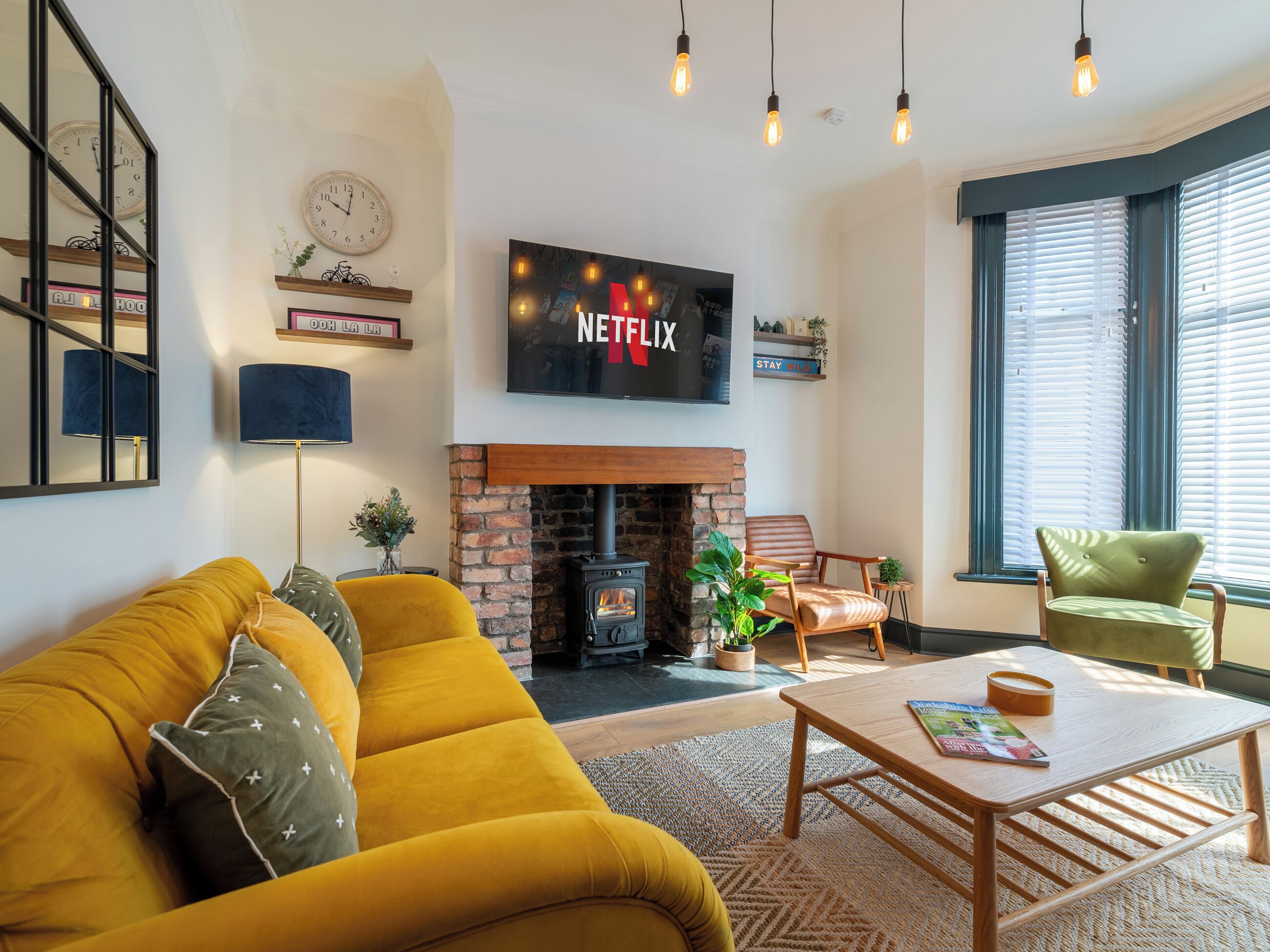 A cozy living room with a yellow sofa, a fireplace, a TV mounted above it, and a large window with blinds.