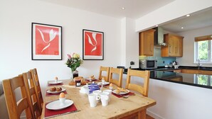 Un comedor con una mesa de madera dispuesta para el desayuno, que incluye cruasanes, huevos y tazas de café. En la pared hay dos cuadros enmarcados, un jarrón con flores y armarios de madera en la cocina contigua.