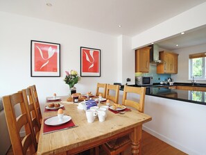 A dining area with a wooden table set for breakfast, including croissants, eggs, and coffee cups. There are two framed artworks on the wall, a vase with flowers, and wooden cabinets in the adjacent kitchen area. - Goosander Lake House (Cotswold Lakes)