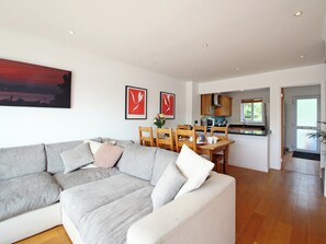 A modern living room with a grey sectional sofa, a dining area with wooden chairs, and a kitchen with wooden cabinets. - Goosander Lake House (Cotswold Lakes)