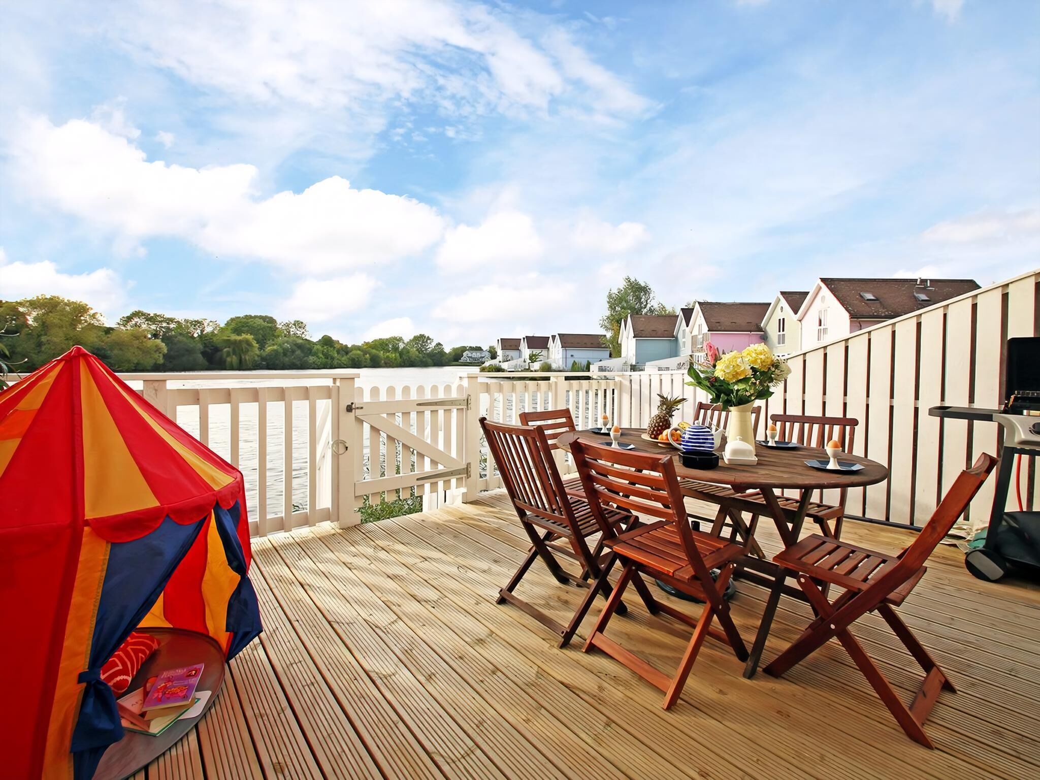 A wooden deck with a table and chairs, a colorful umbrella, and a view of houses across a body of water.