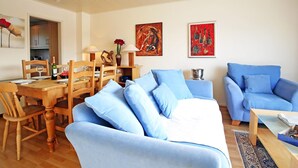 Uma sala de estar com uma mesa de jantar de madeira, sofá azul e duas pinturas na parede.