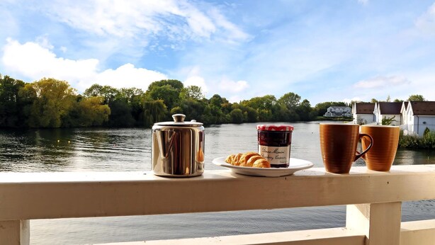 Vista ribeirinha com um deck de madeira, um contêiner metálico, um prato com croissants e geleia, e duas canecas.