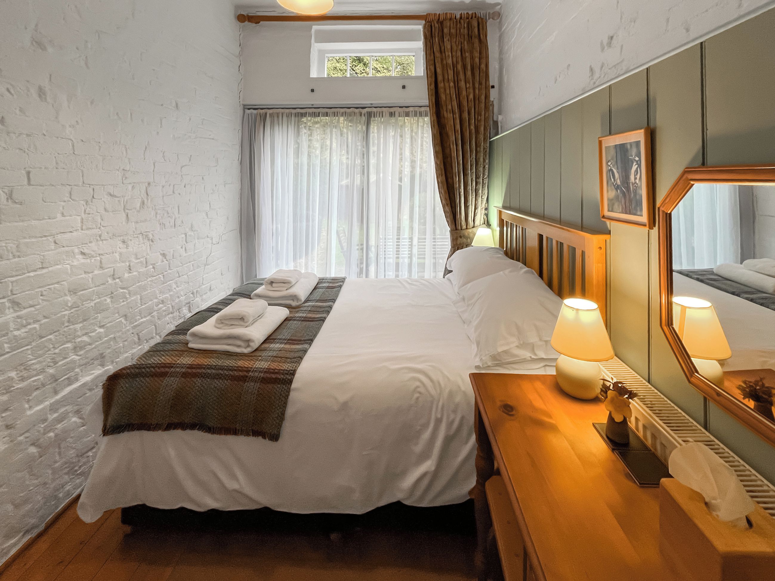 Habitación de hotel con cama, mesita de noche con lámpara, espejo y ventana con cortinas.