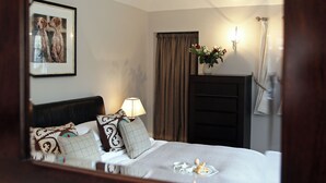 Ein Hotelzimmer mit einem Bett, einem gerahmten Bild an der Wand, einer Nachttischlampe, einer Vase mit Blumen und einem Spiegel, der den Raum reflektiert.