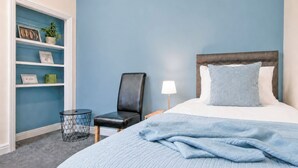 Un dormitorio con cama, una silla, una mesita de noche, una lámpara y una estantería con objetos decorativos.