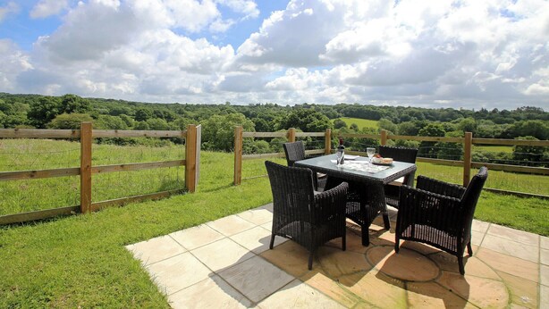Eine Terrasse im Freien mit Tisch und Stühlen, einer Weinflasche und einem Glas, vor einer grünen Wiese und einem Holenzaun.