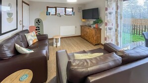 Ein Wohnzimmer mit zwei braunen Ledersofas, einem kleinen Holztisch, einem Fernseher auf einer Holzkommode und Blick ins Freie durch ein Fenster mit Vorhängen.