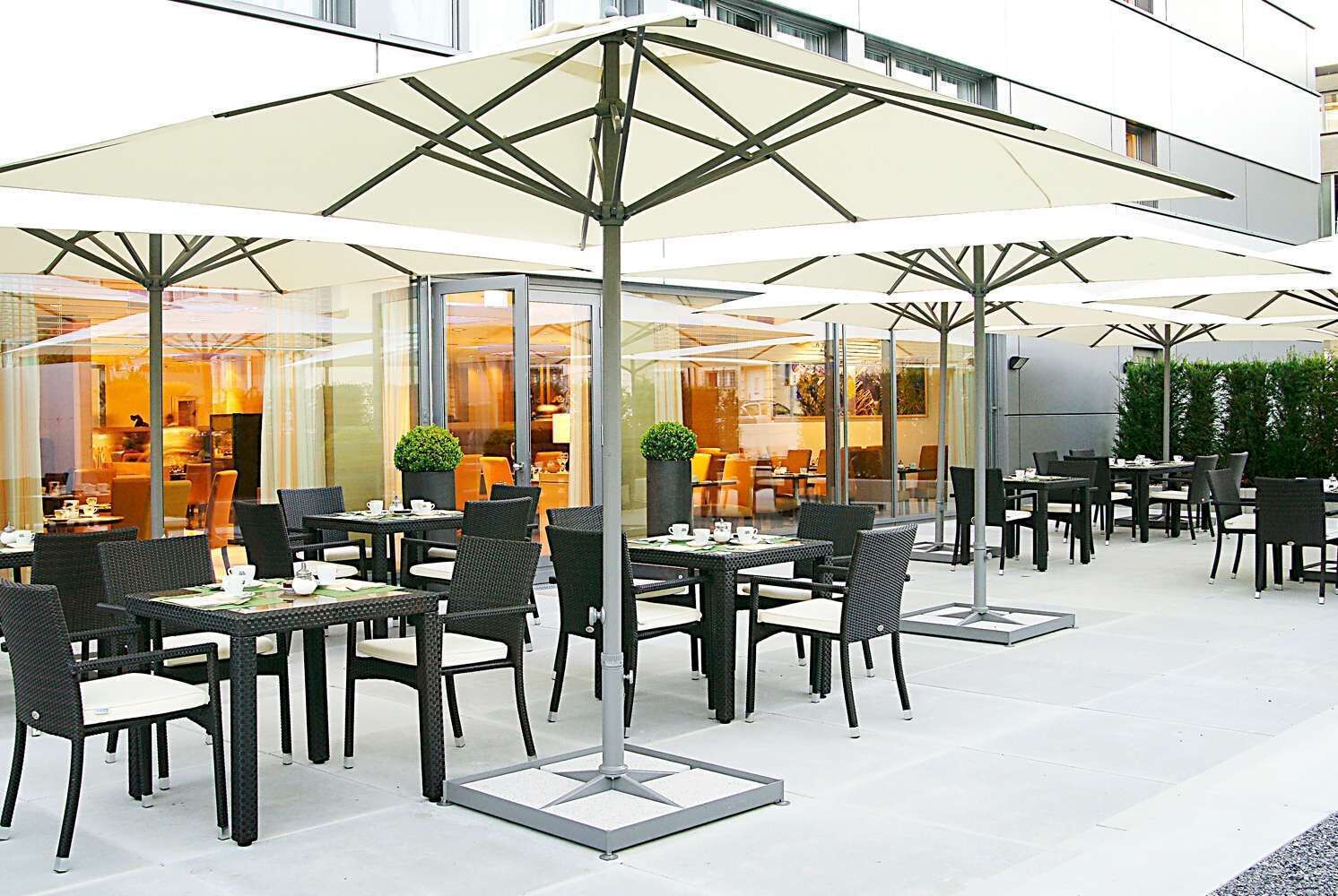 Un'area caffè all'aperto con tavoli e sedie, grandi ombrelloni e una parete di vetro che offre vista sull'interno.