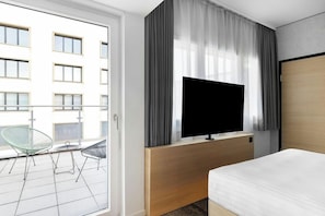 A hotel room with a bed, a TV mounted on a wooden console, a balcony with a chair, and a view of a building with multiple windows. - H Hotel Zurich (Zürich)