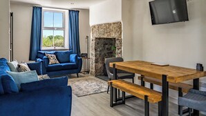 A living room with a blue sofa, a wooden dining table with benches, a stone fireplace, and a flat-screen TV mounted on the wall. - 3 bedroom accommodation in Beaumaris (Beaumaris)