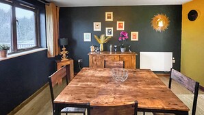 Una sala da pranzo con un tavolo e sedie in legno, una finestra vista alberi e un muro con quadri e un decoro solare.