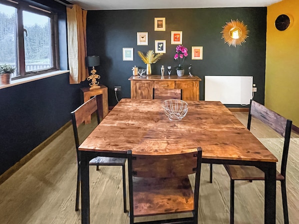 A dining room with a wooden table and chairs, a window with a view of trees, and a wall with framed pictures and a sun decoration.
