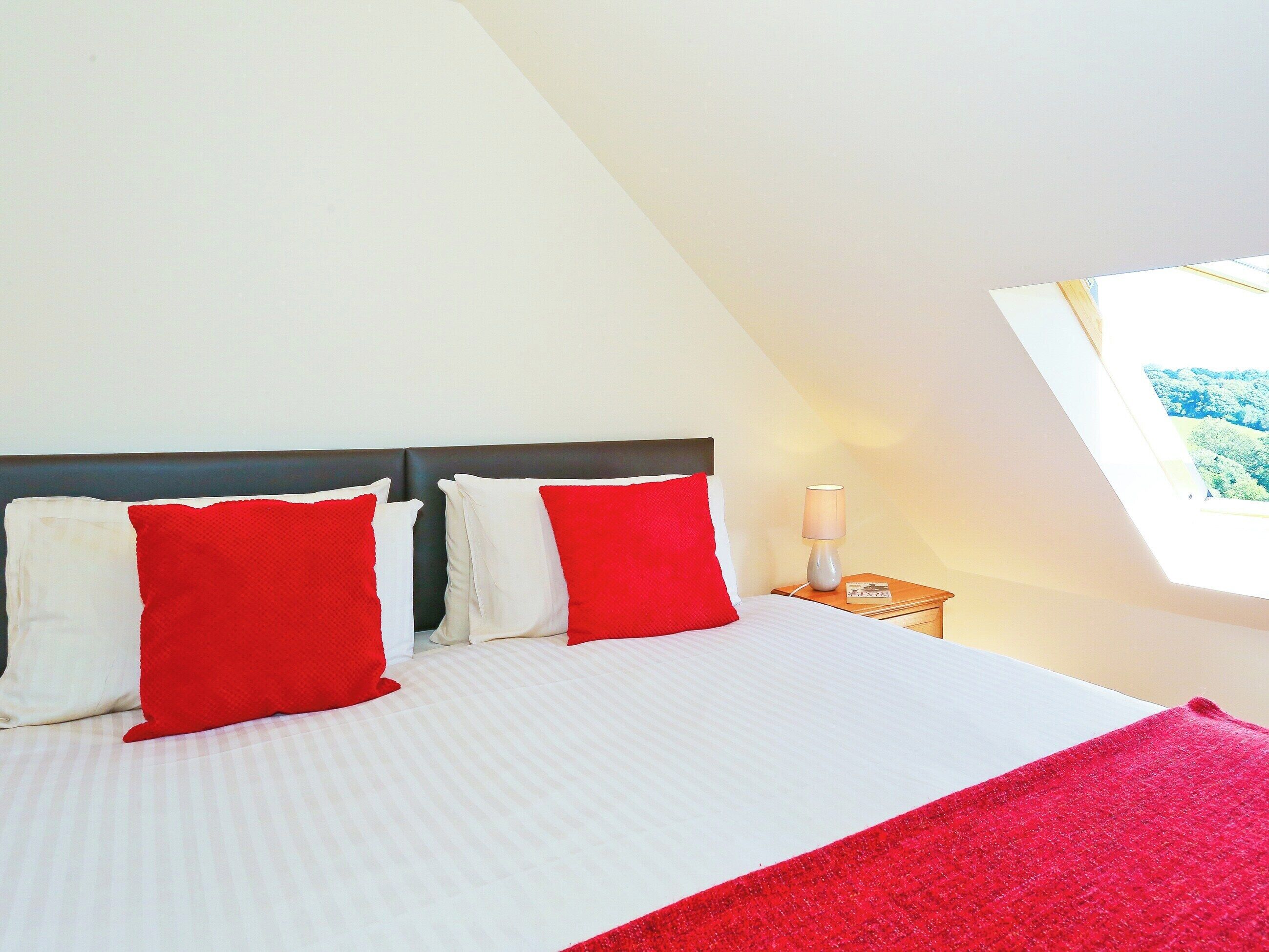 Ein Schlafzimmer mit einem Bett, zwei roten Kissen, einem Nachttisch mit Lampe und einem Fenster mit Blick.