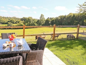 A table with a fruit bowl, cups, and a teapot, set against a backdrop of a grassy field with a wooden fence and sheep grazing.