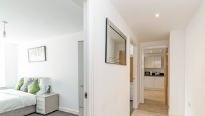 Un dormitorio moderno con cama, mesita de noche y espejo. Un pasillo conduce a una zona de cocina con armarios y microondas.
