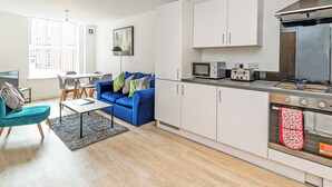 Una cocina moderna con gabinetes blancos, microondas, tostadora y horno. En la zona de estar contigua hay un sofá azul y una mesa de comedor con sillas.