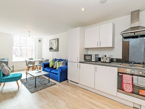 A modern kitchen with white cabinets, a microwave, toaster, and oven. There is a blue sofa and a dining table with chairs in the adjacent living area. - Apartment 30 - UK42613 (Preston)