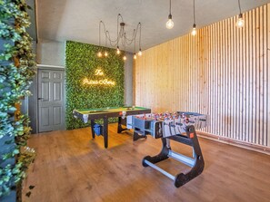 A room with a pool table, foosball table, and a green wall with a neon sign.