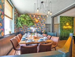 En matplats med ett långt bord dukat för en måltid, omgivet av stolar, och en vägg dekorerad med ett blommönster samt en grön, vertikal trädgård.