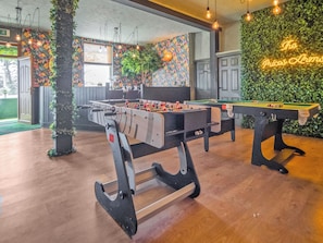 Une table de billard, une table de baby-foot et une table de flipper dans une pièce au plancher en bois et au mur vert orné d’un néon.