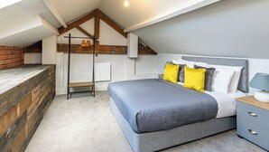 Un dormitorio con techo inclinado, una cama con ropa de cama gris y almohadas amarillas, una mesita de noche con lámpara, y un banco de madera apoyado en la pared.
