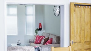 Een woonkamer met een grijze bank, rode en witte kussens, een houten deur en een klok aan de muur.
