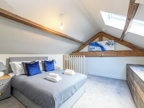 Un dormitorio con techo inclinado, una cama con ropa de cama gris y almohadas azules, una mesita de noche con lámpara y un cuadro en la pared.