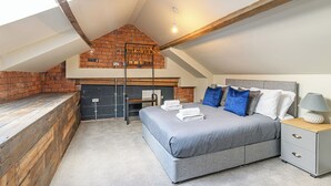 A bedroom with a sloped ceiling, exposed brick wall, a bed with grey bedding, a bedside table with a lamp, and a hanging light fixture.