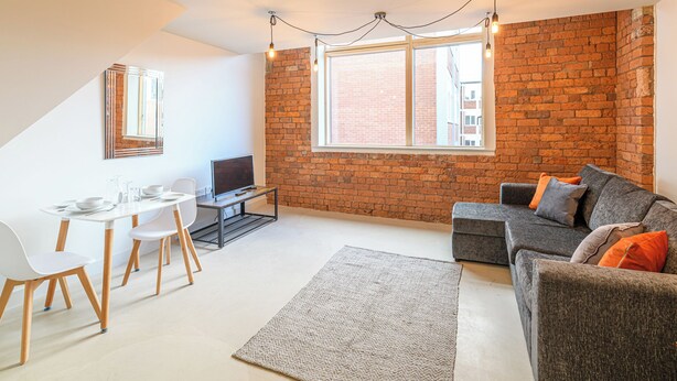 A modern living room with a brick wall, a grey sofa with orange cushions, a white dining table with chairs, and a TV on a stand.