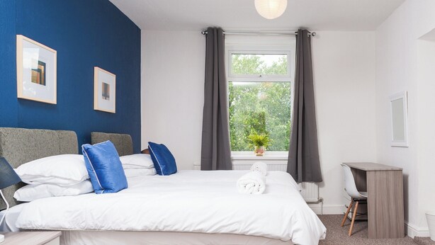 A bedroom with a bed, bedside tables, a desk, and a window with curtains. - Newport House Crindau Road Upper (Newport)